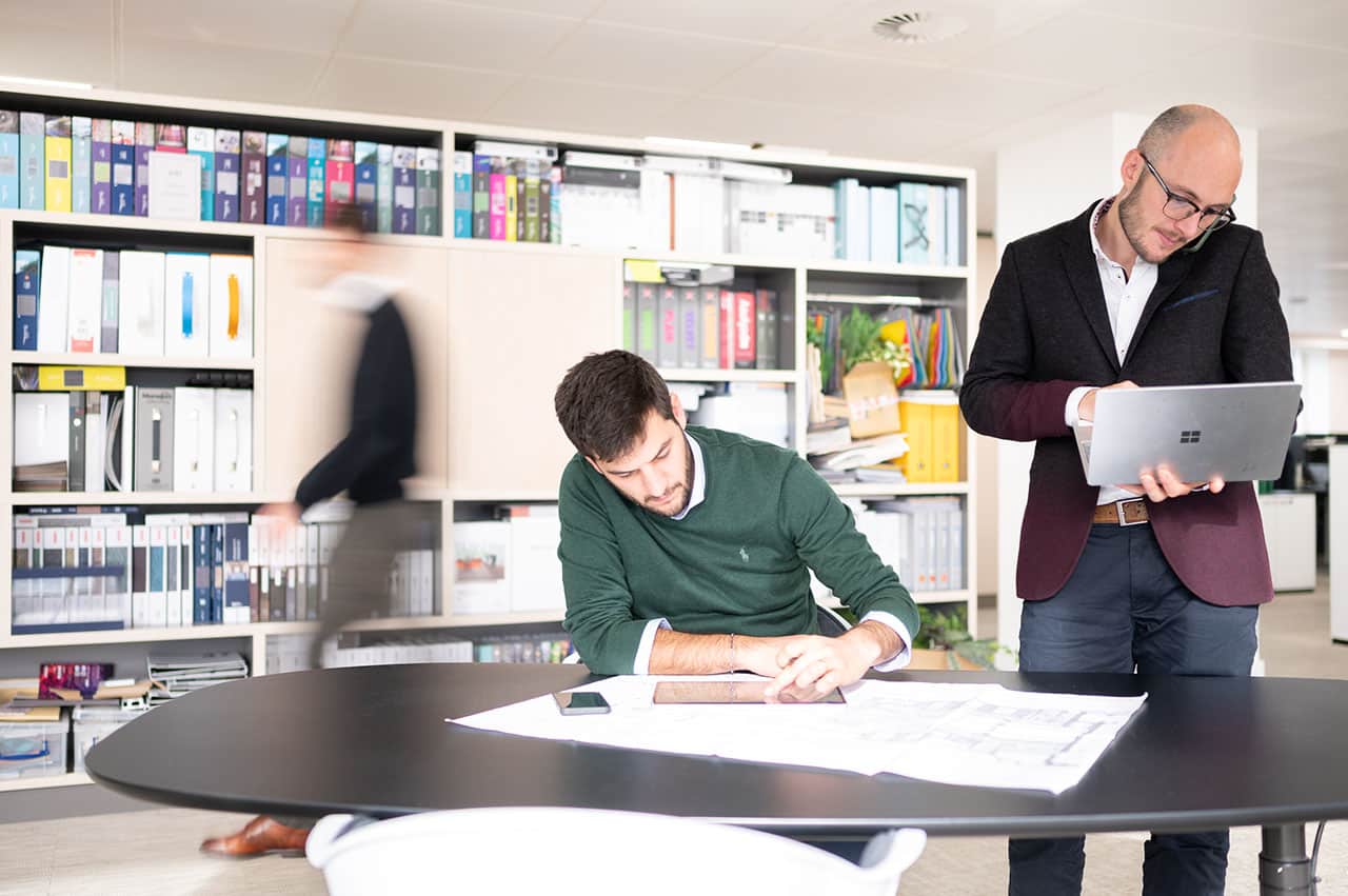 Man draws at a table in an office while his colleague works on his laptop