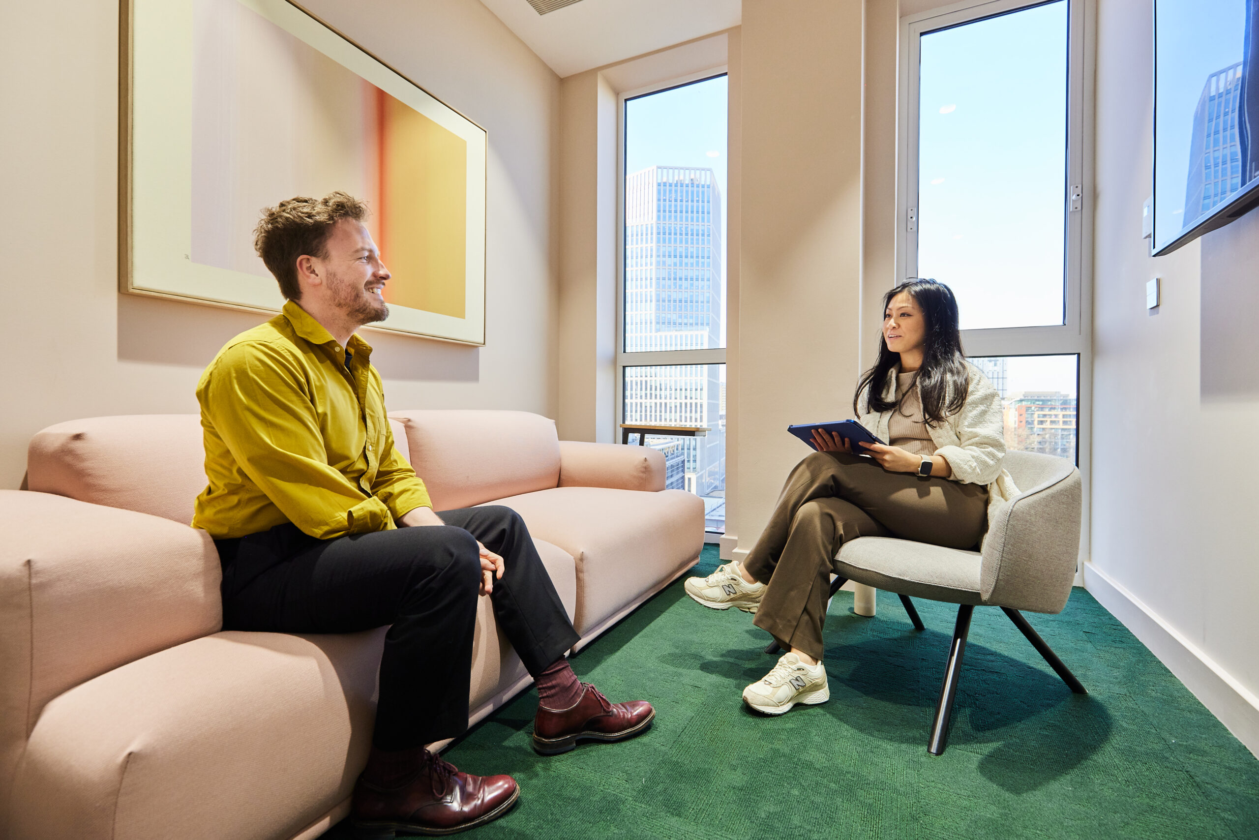 Two people seated in a modern office meeting room, having a conversation during a one-to-one discussion, with city buildings visible through large windows.